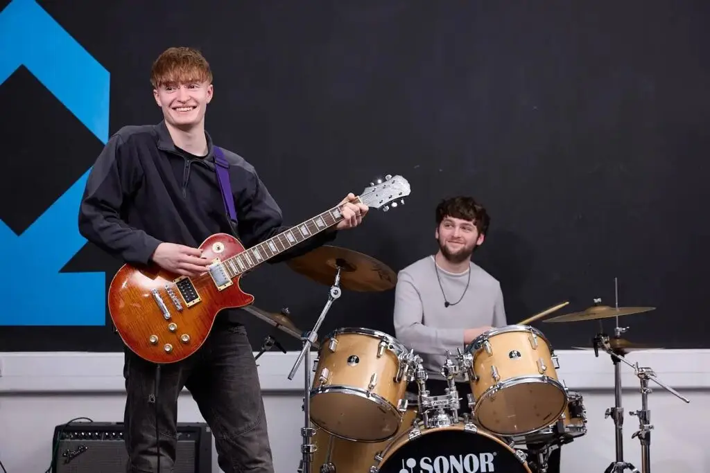 Two students on stage with a guitar and drums, smiling and ready for a performance.
