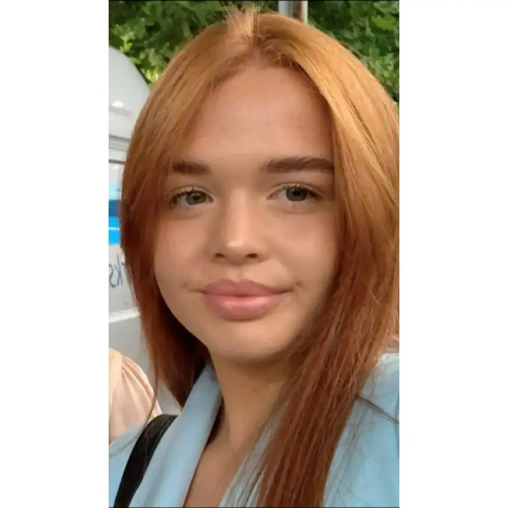 A young woman with long red hair and a light blue top smiles at the camera, standing outdoors with greenery in the background.