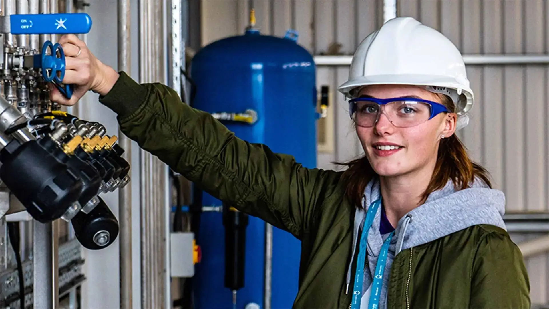 A woman in a hard hat and safety glasses adjusts a blue valve in an industrial setting, showcasing practical engineering skills.