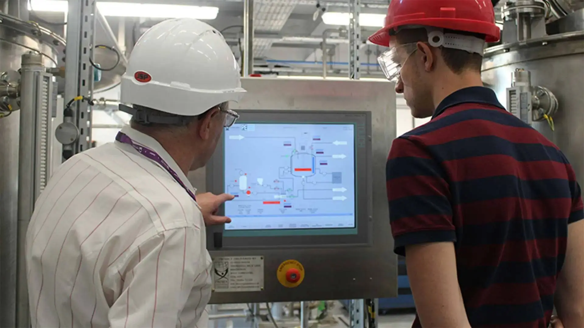 Two men in hard hats and safety glasses examining a control panel with diagrams in an industrial setting.