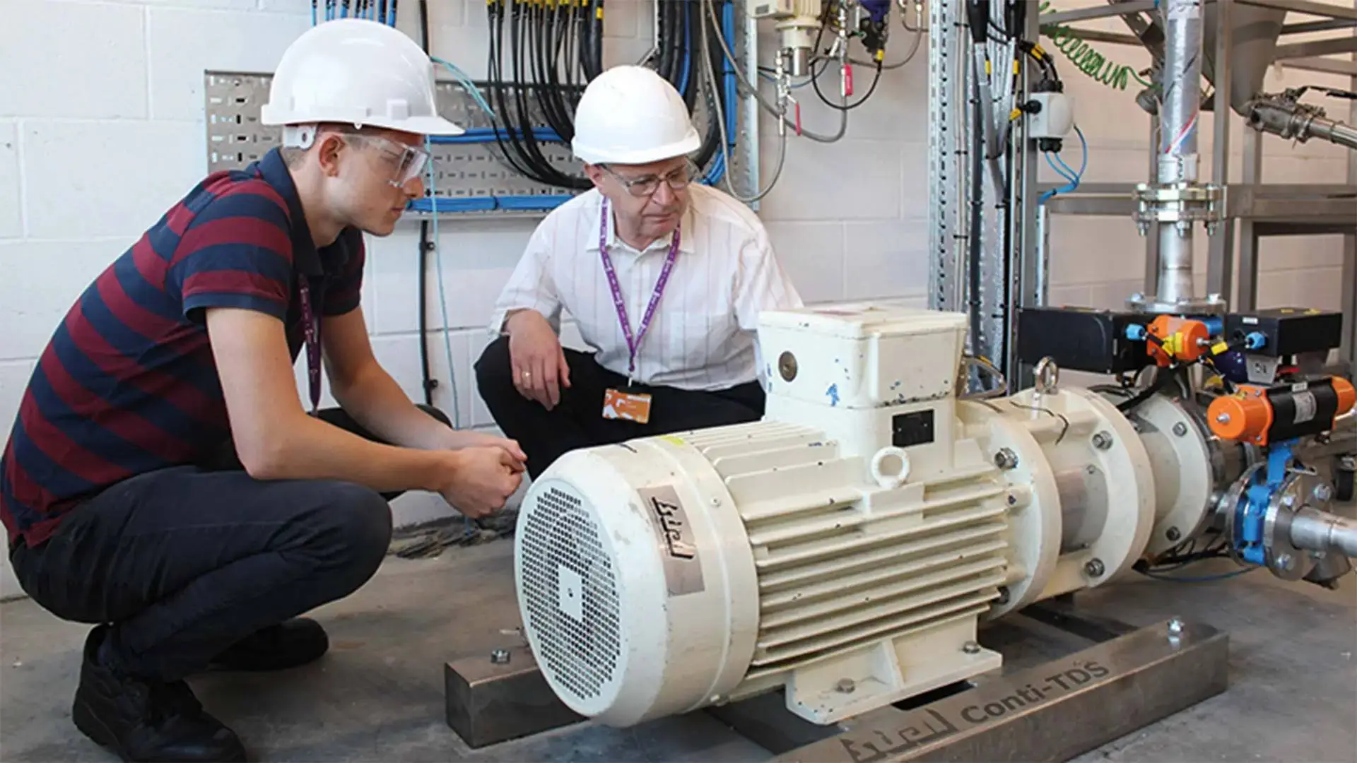 Two individuals in hard hats and safety glasses examining a large electric motor in a technical workshop setting.
