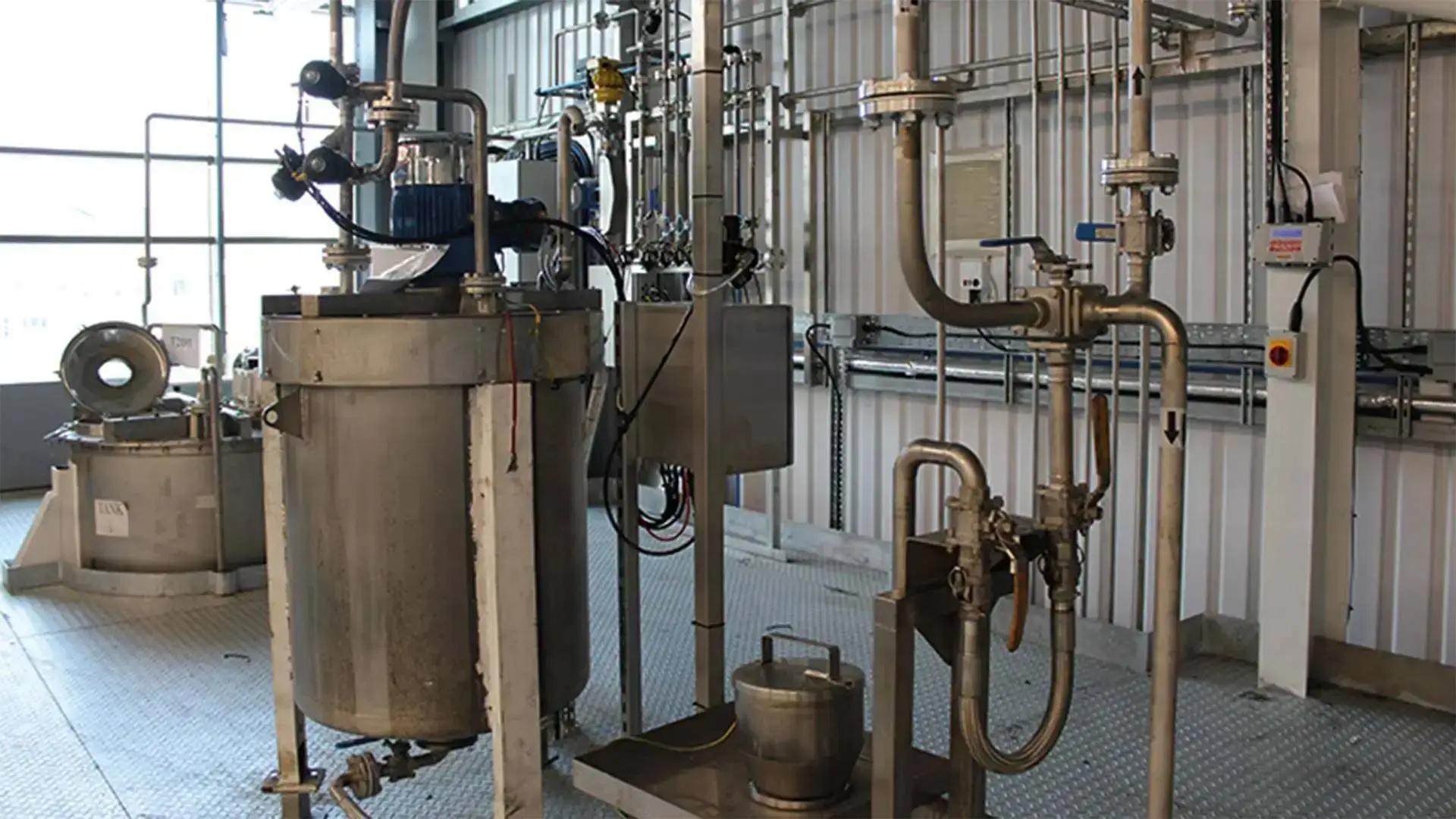 Stainless steel industrial tanks and pipes in a well-lit room, likely part of a chemical or food processing facility, with control panels and safety features.