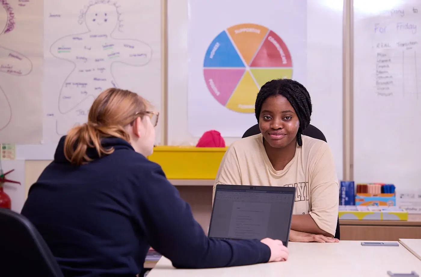 Two people sitting at a table with a laptop, engaged in conversation. Educational posters and a pie chart on the wall behind them.