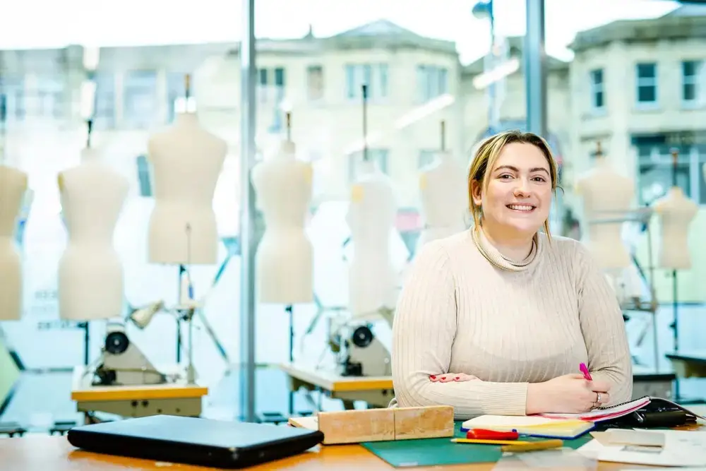 A smiling student in a beige sweater sits at a desk with notebooks and a tablet, surrounded by dress forms in a fashion design studio.