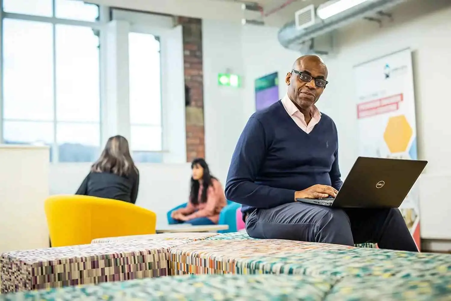 A man in a navy jumper using a laptop on a colourful bench in a college lounge with students in the background.