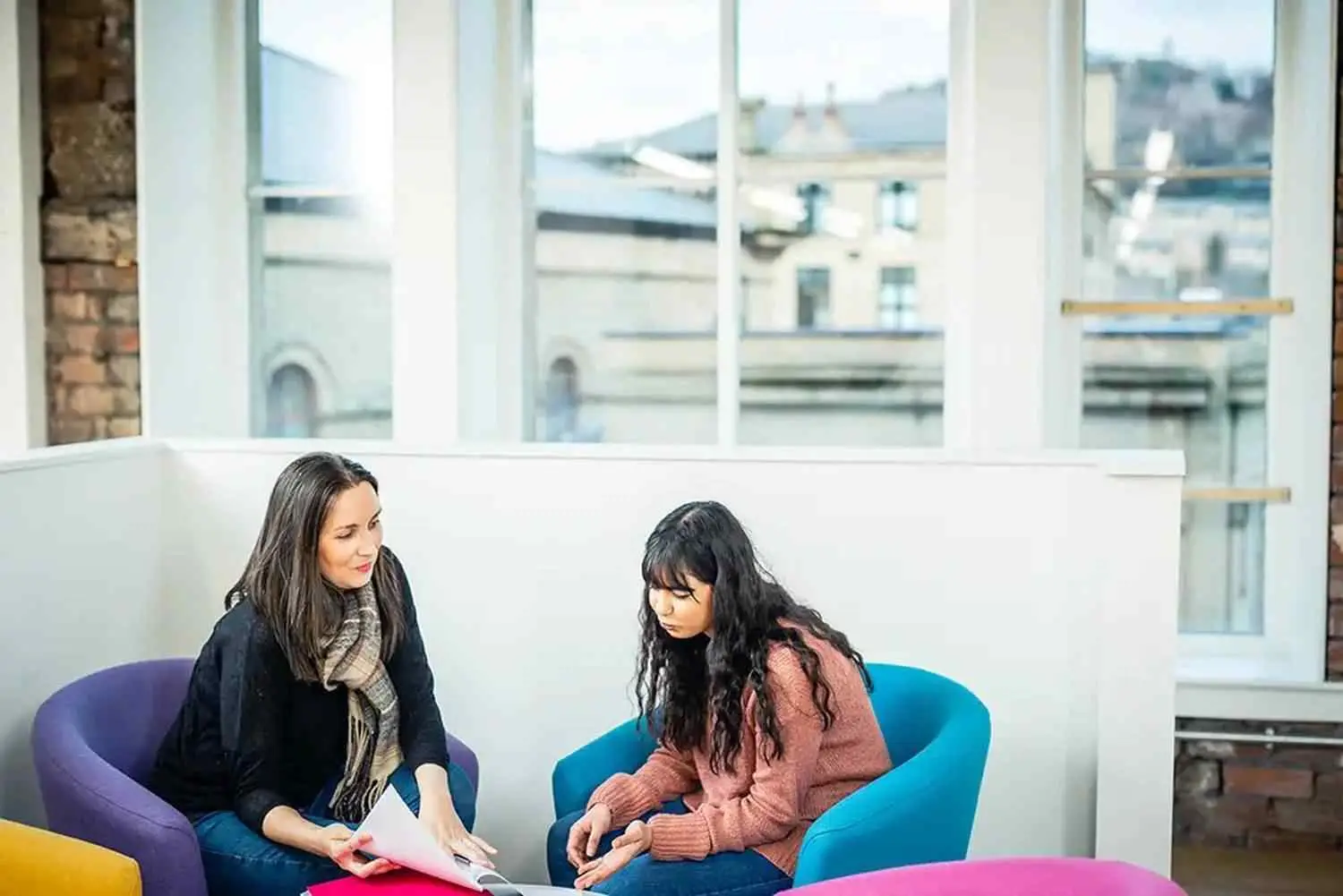 Two students in a college lounge, seated on colourful chairs, discussing documents and a laptop. Large windows show a cityscape.