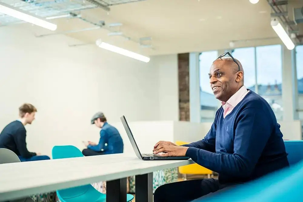 man in a blue sweater smiling and typing on a laptop at a white table in a bright college study area.