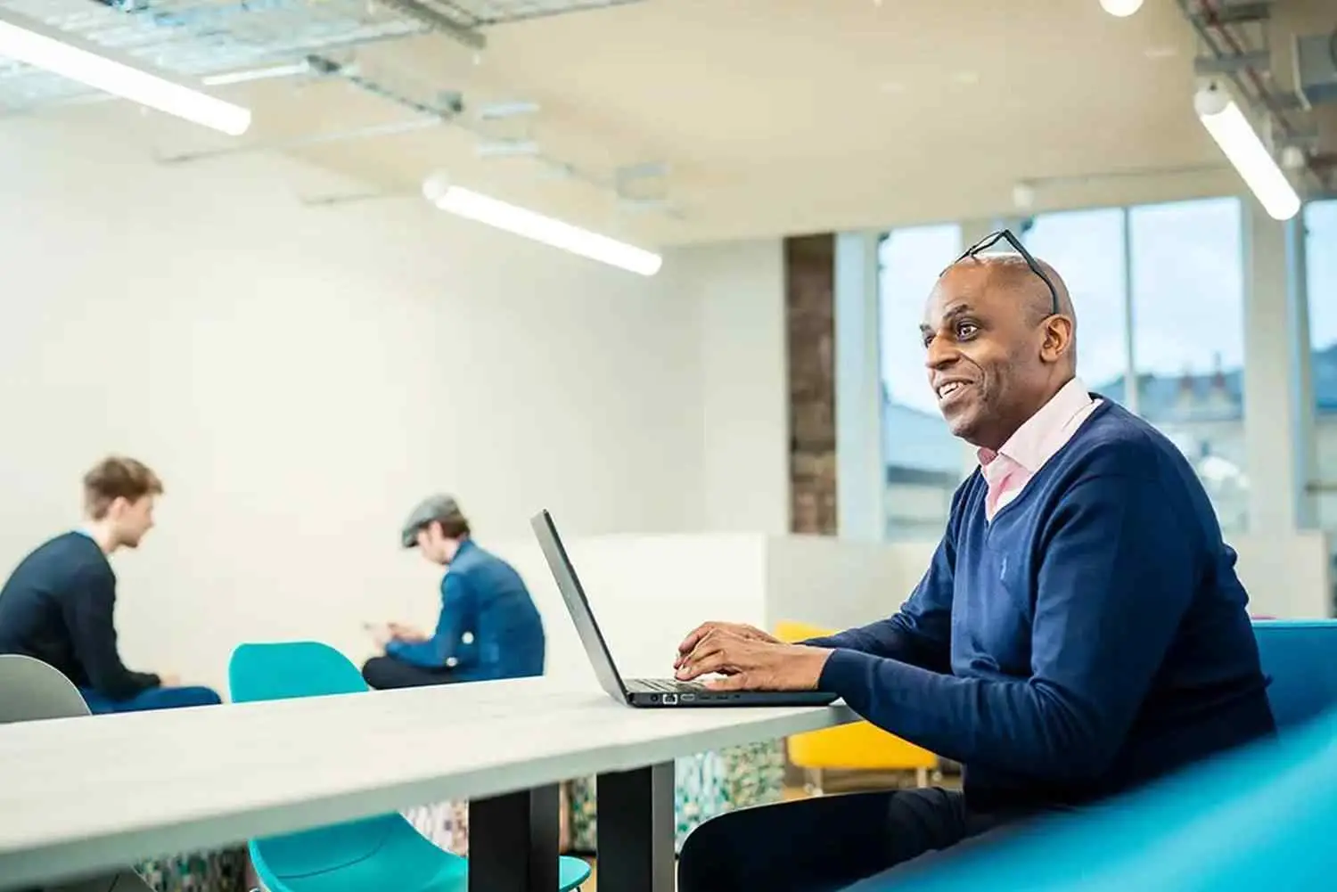 A man in a blue sweater working on a laptop in a bright, modern college library with other students studying in the background.