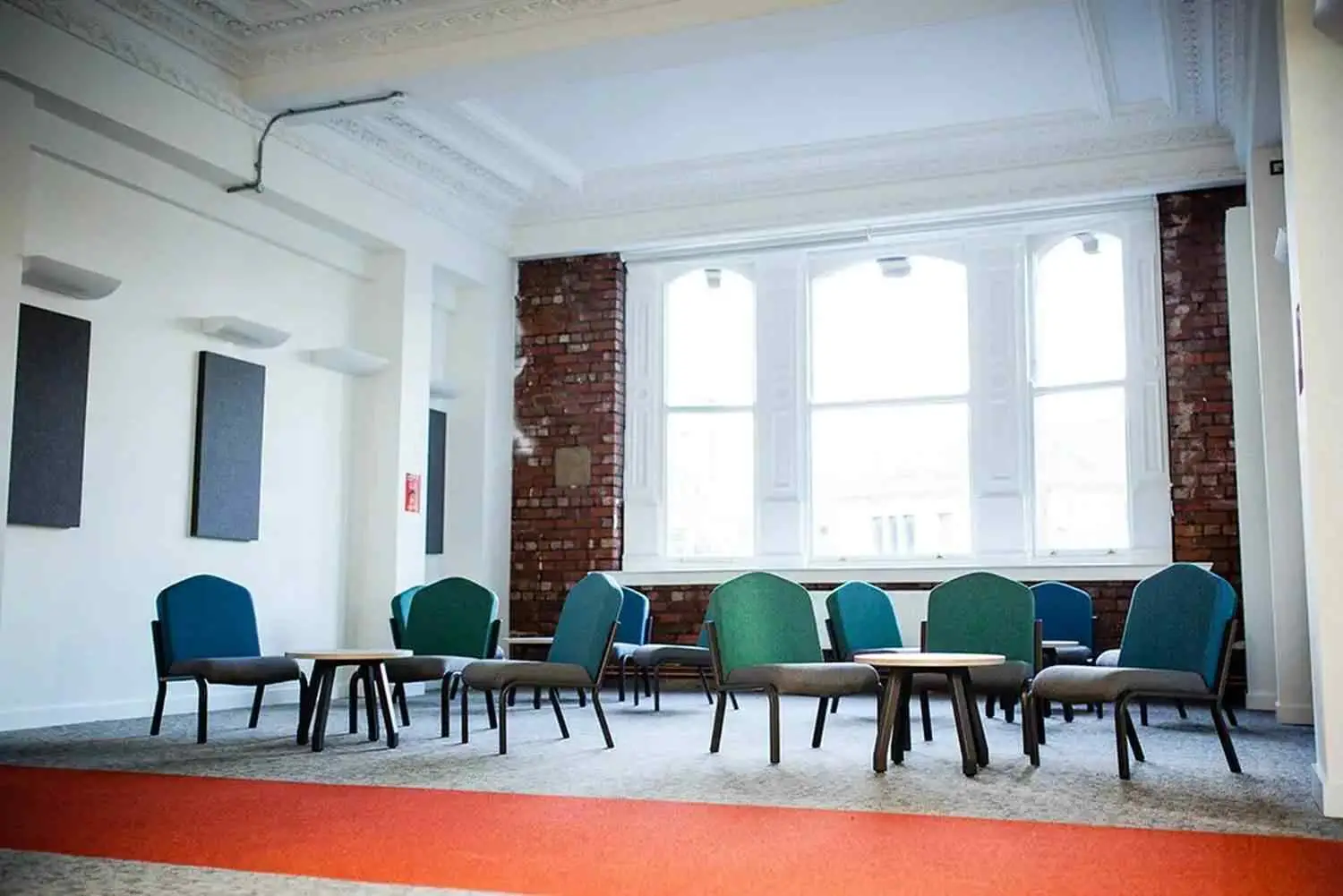 A bright classroom with green chairs, wooden tables, and exposed brick walls. Large windows let in natural light, creating an inviting learning environment.