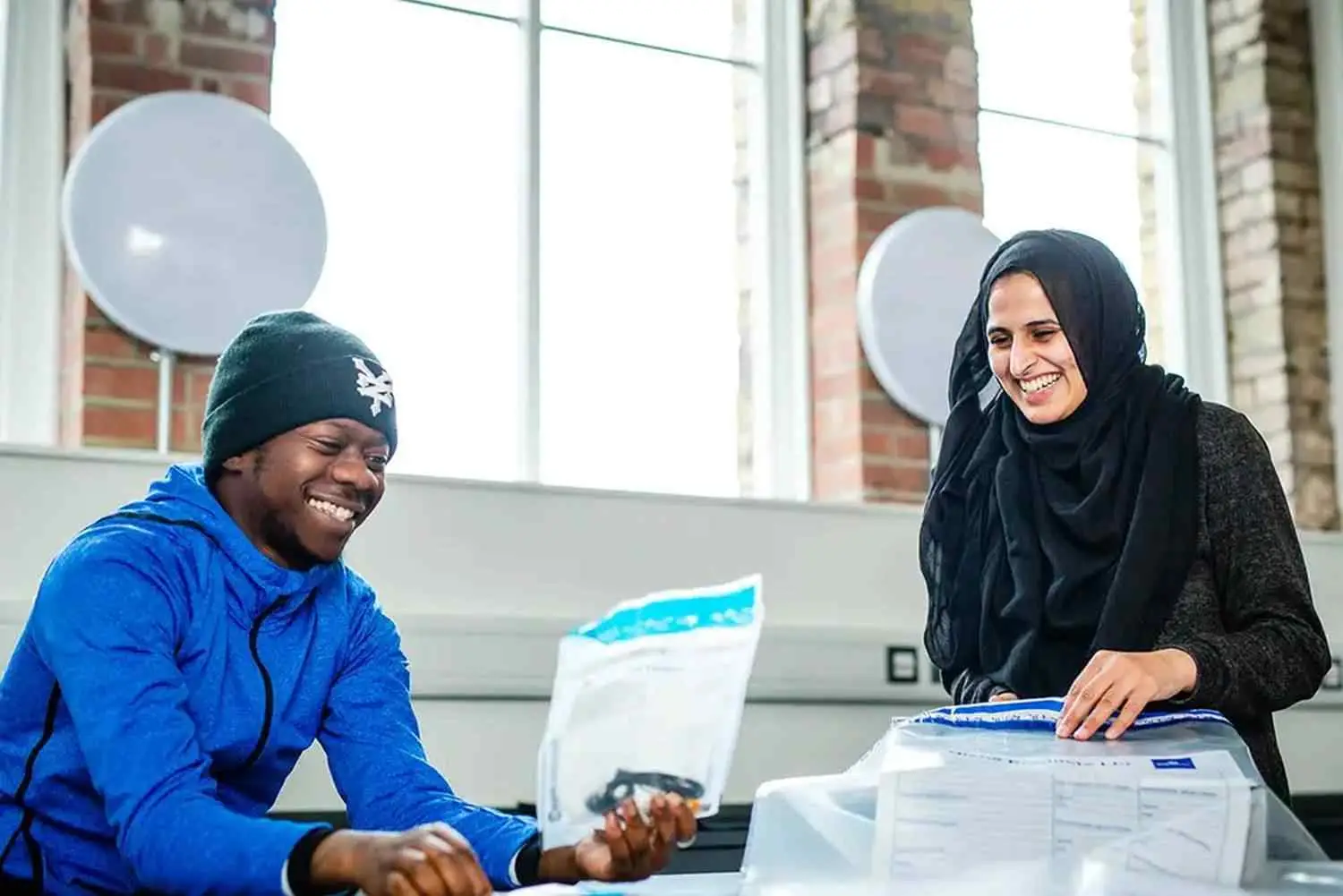 Two students smiling and shaking hands at a desk with papers and envelopes in a bright room with brick walls and large windows.