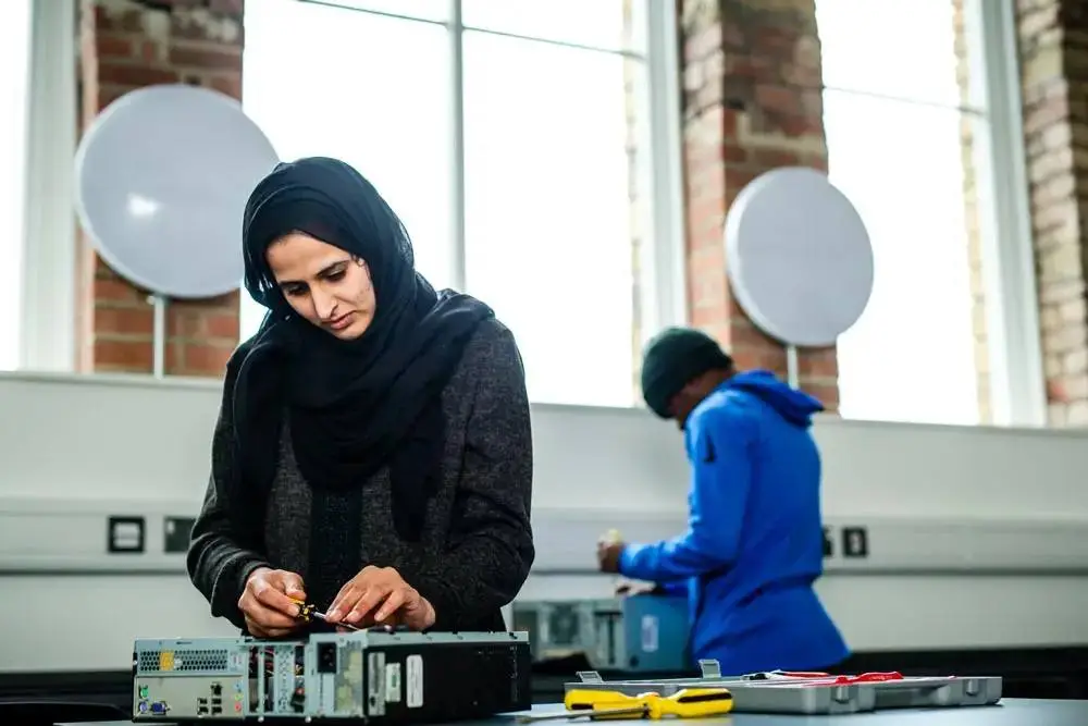 Two students in a tech lab, one focused on assembling a computer, surrounded by tools and equipment, under large windows with industrial brick walls.