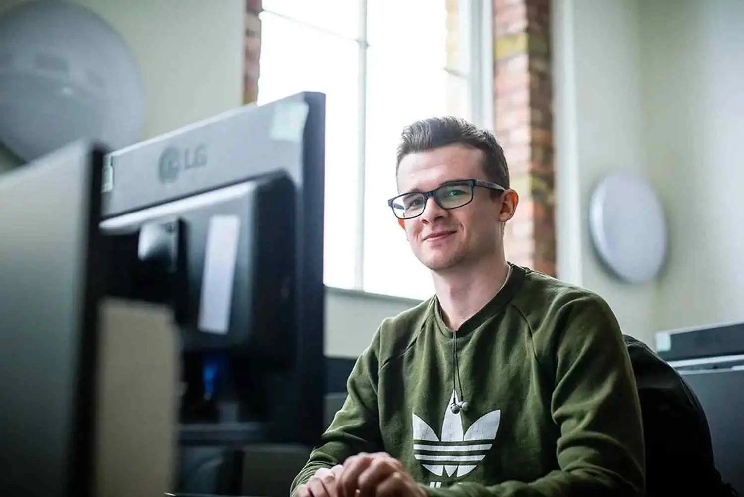 A man in glasses sits at a desk, smiling at a computer screen in a brightly lit room with brick walls.