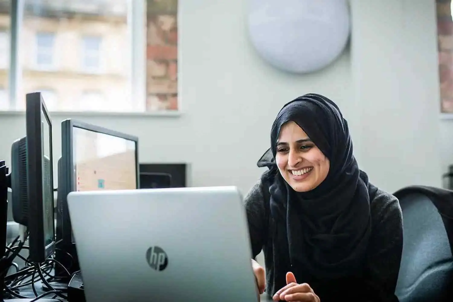 A woman in a black top smiling at a laptop in a brightly lit room with a window and a clock on the wall.