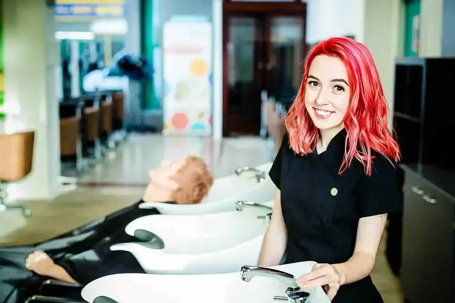 A woman with red hair stands next to a salon sink with a mannequin head, smiling at the camera.