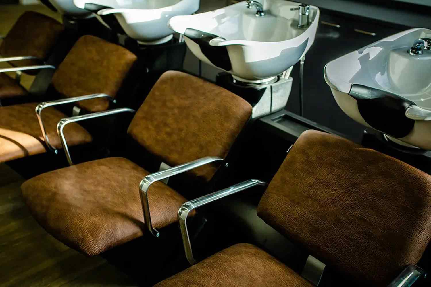 A row of brown salon chairs with chrome armrests in front of white sinks, likely in a hairdressing training facility.