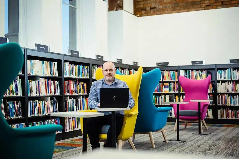 man using a laptop in a library, seated in a yellow chair between teal and pink chairs, with bookshelves in the background.