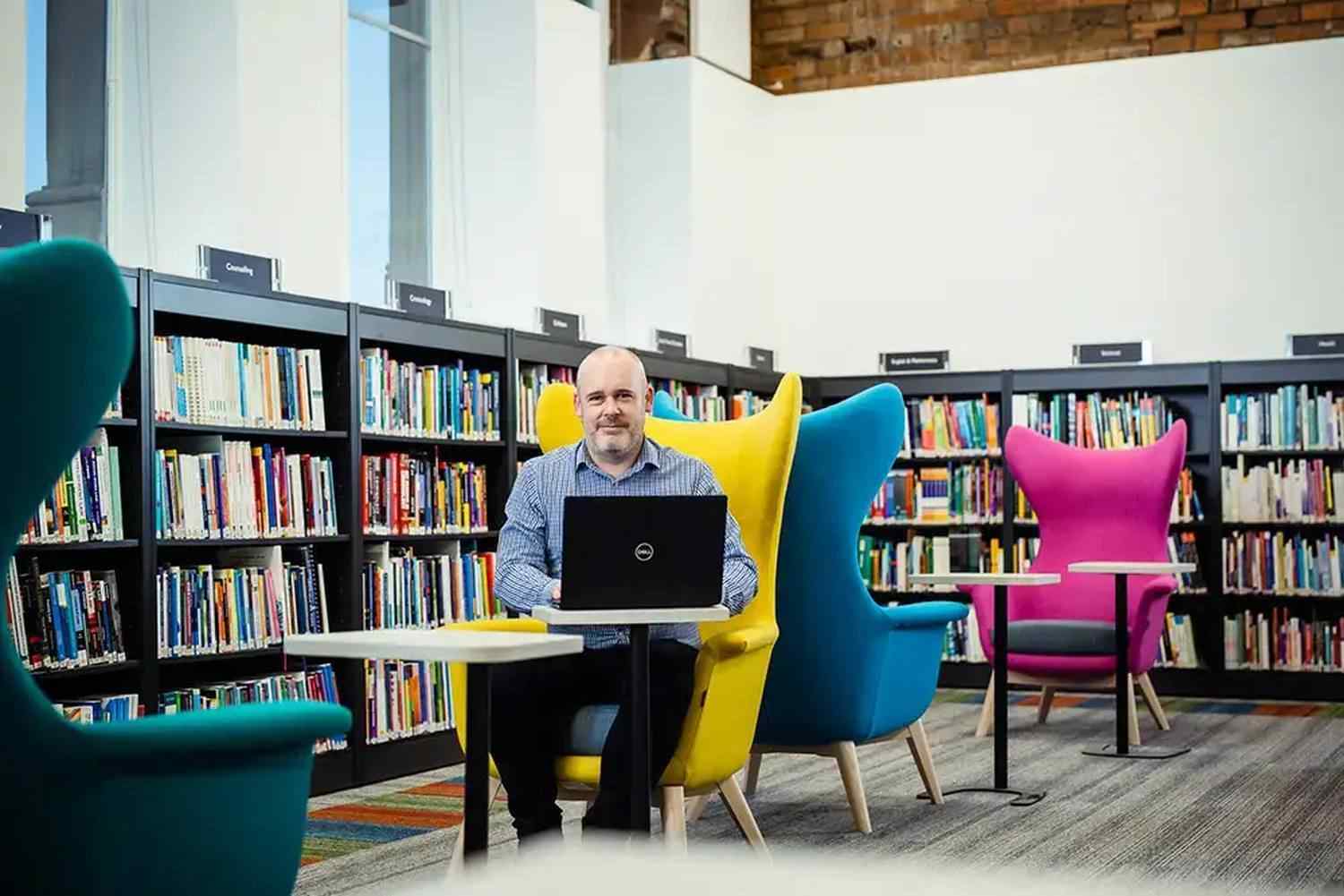 A man using a laptop in a library, seated between colourful chairs. Bookshelves line the background.