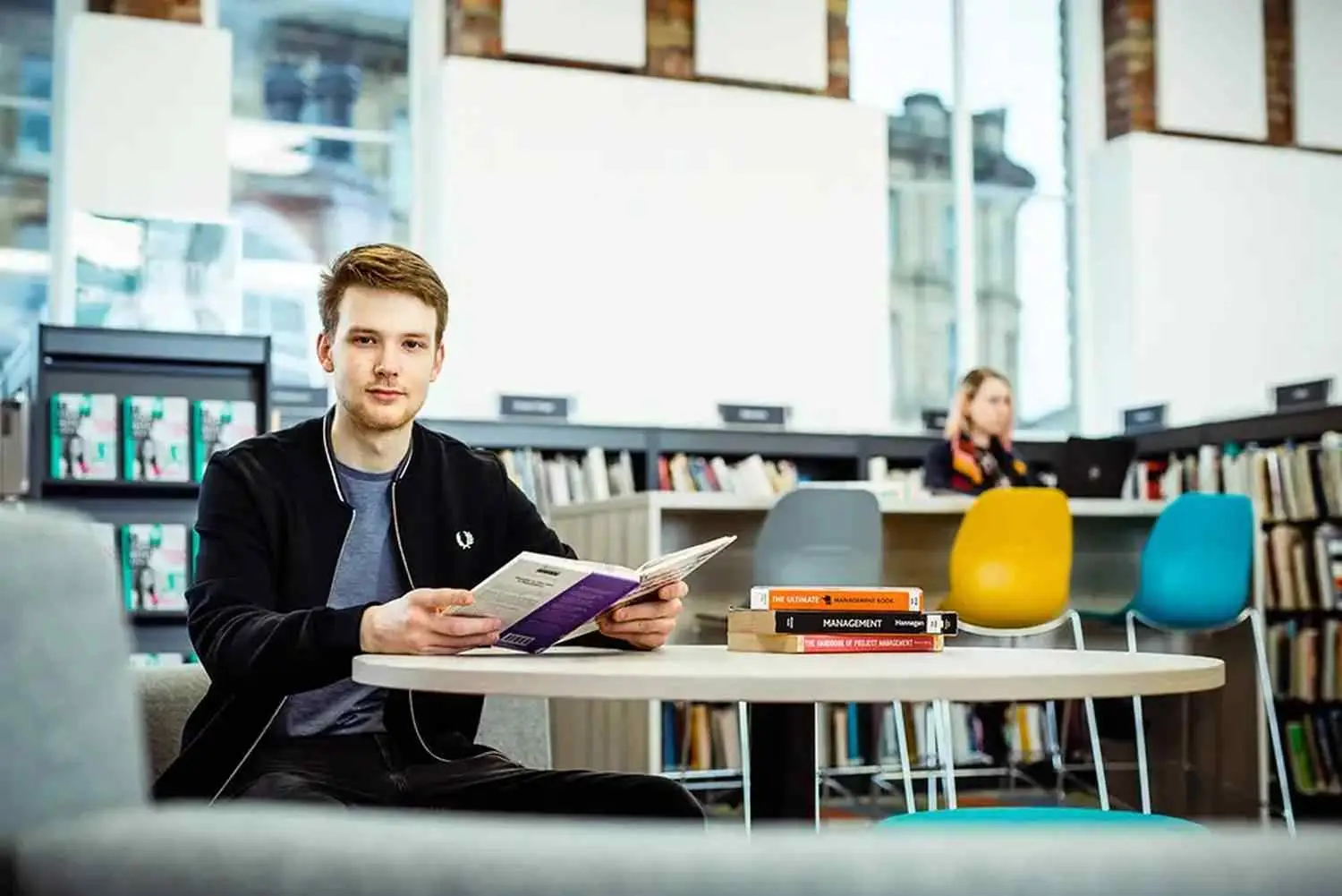 A student in a library reads a brochure at a table with textbooks. Bright, modern setting with bookshelves in the background.