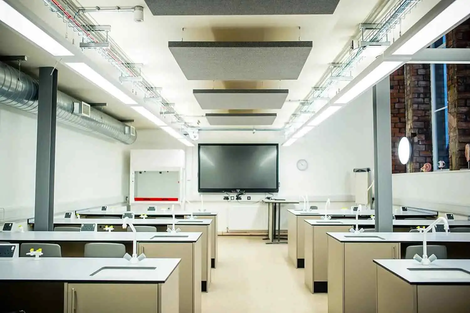 A modern college science lab with white benches, sinks, and a large screen at the front. Exposed brick wall and industrial lighting create a contemporary learning environment.