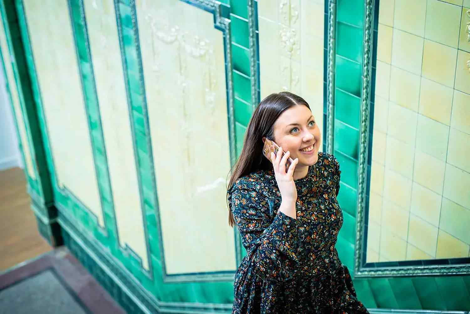 A woman in a floral dress smiling and talking on a mobile phone in a tiled corridor.