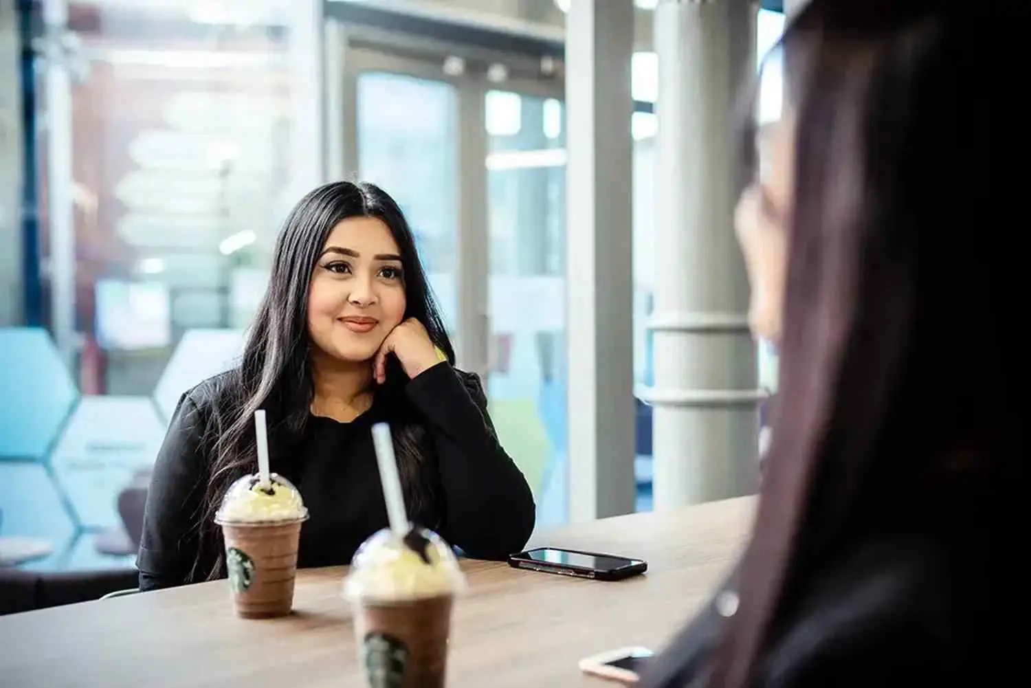 Two women sitting at a table with drinks, engaged in conversation in a modern, well-lit college cafe.