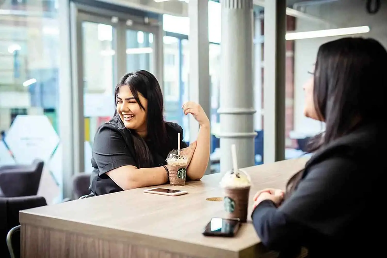 Two students smiling and chatting over iced coffee at a table in a brightly lit college cafe.