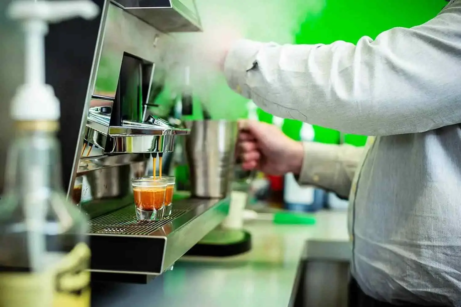 A barista in a white shirt uses an espresso machine to brew coffee into a small glass in a café setting.