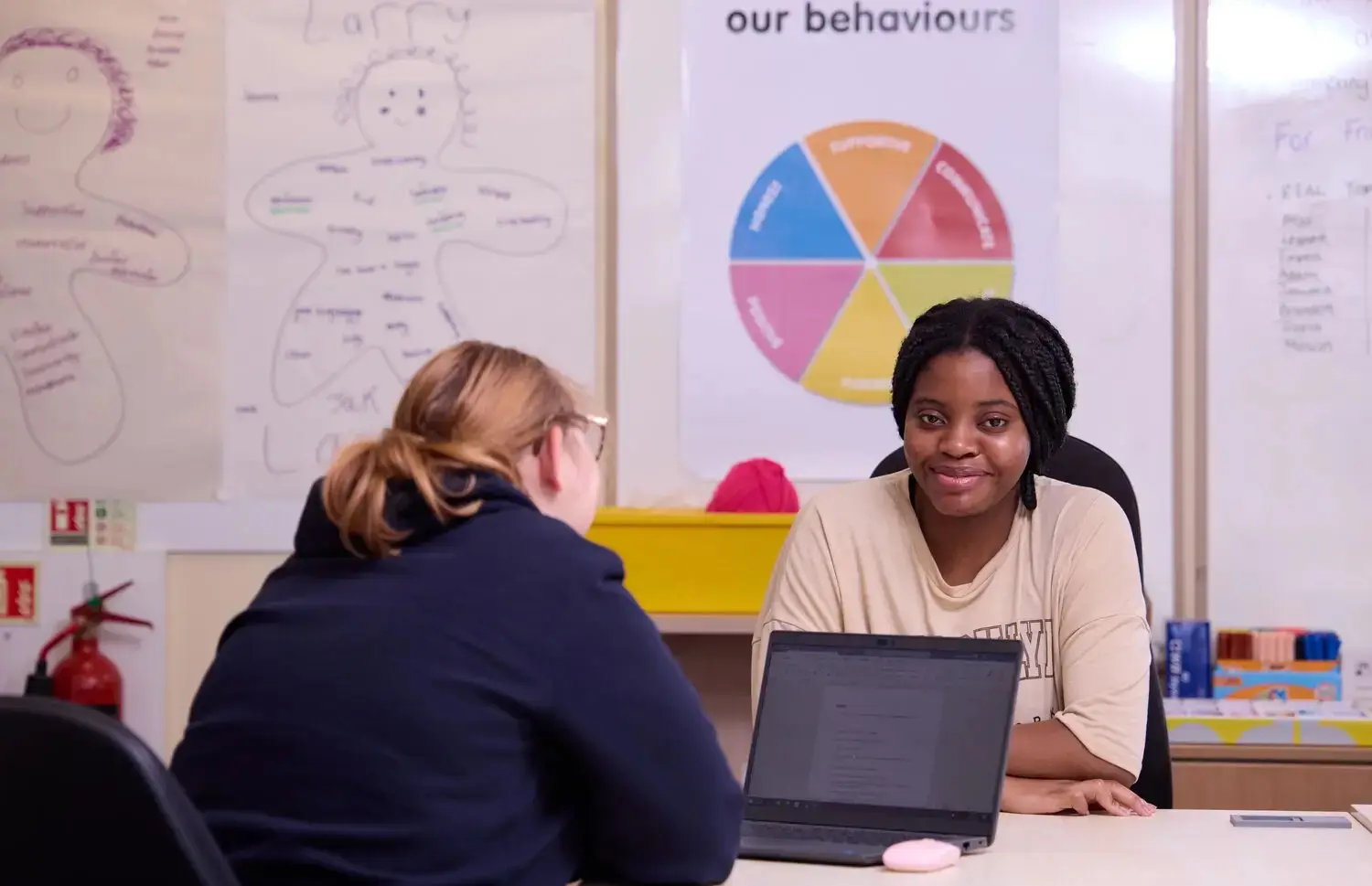 Two people sitting at a table with a laptop, discussing in front of educational posters and a behaviour chart.