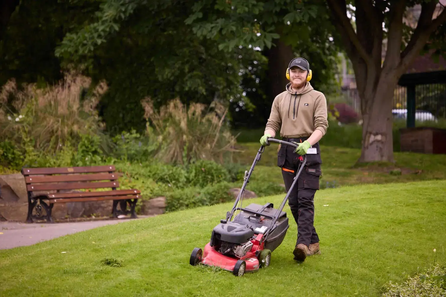 A man in protective gear mowing a lush green lawn in a park, with trees and a bench in the background.