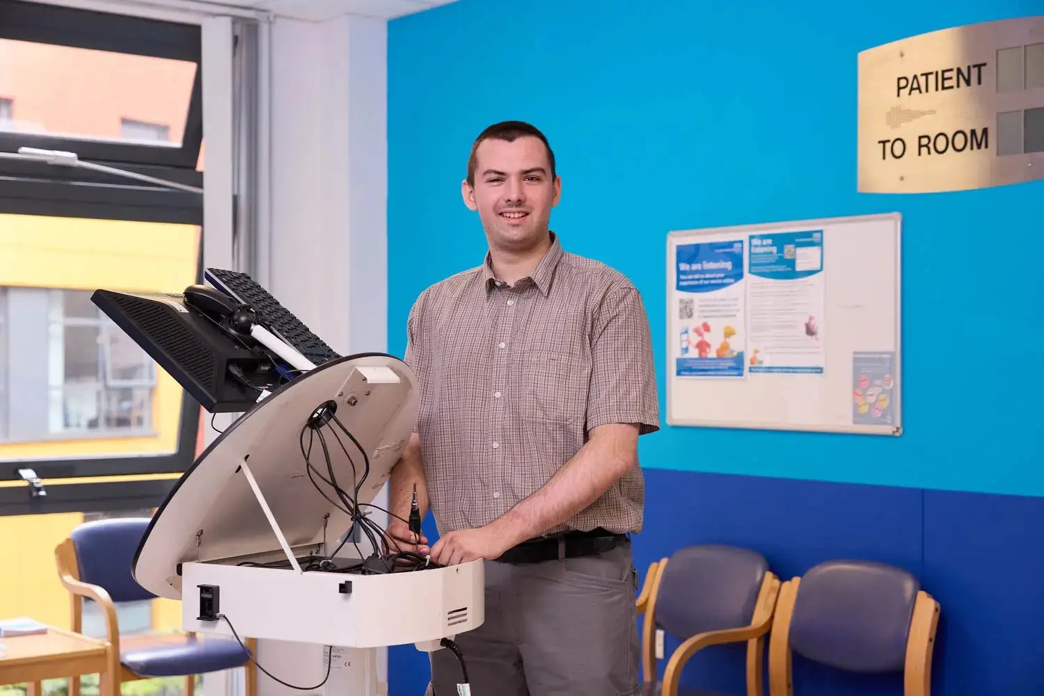 A man in a checkered shirt stands next to a medical device in a brightly coloured waiting room with blue and white walls.