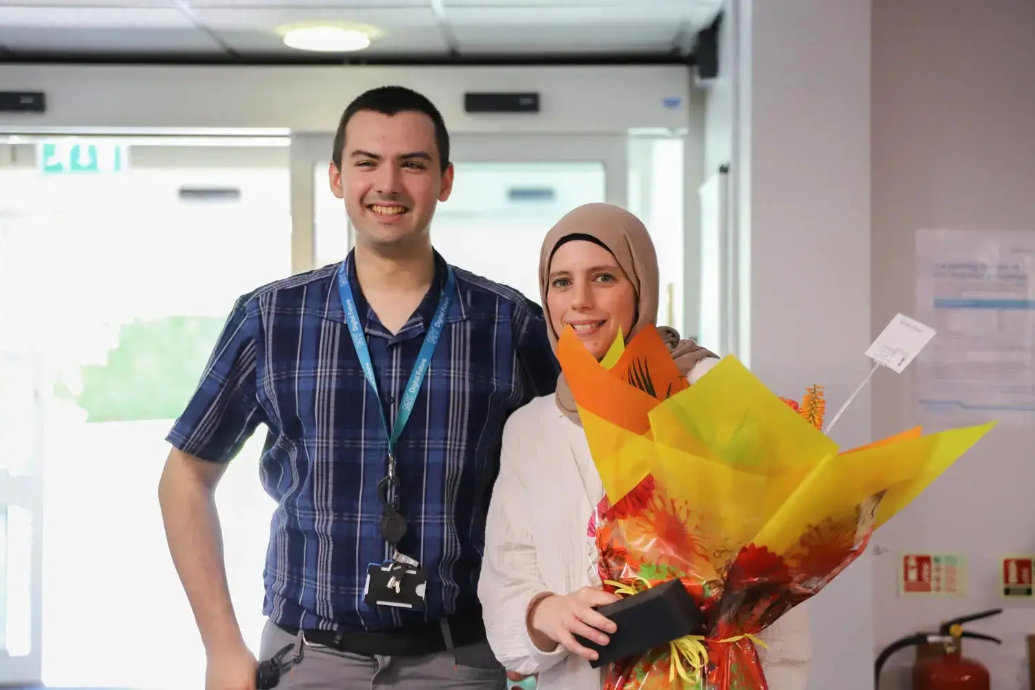 Two people smiling in a college hallway, one holding a colourful bouquet and a certificate.