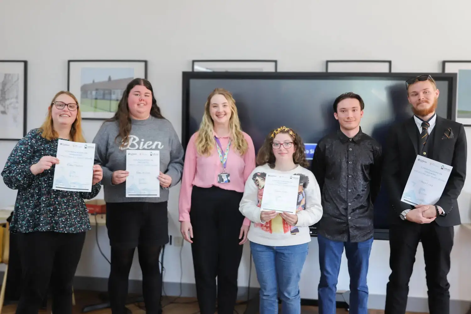 Group of six students holding certificates in front of a screen in a classroom.