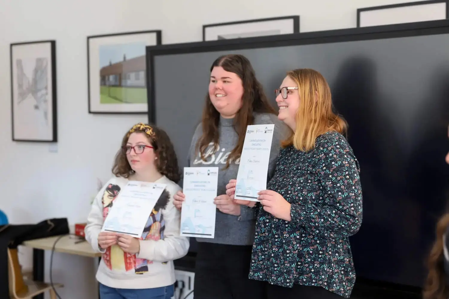 Three students holding certificates in a classroom, smiling and standing in front of a blackboard.