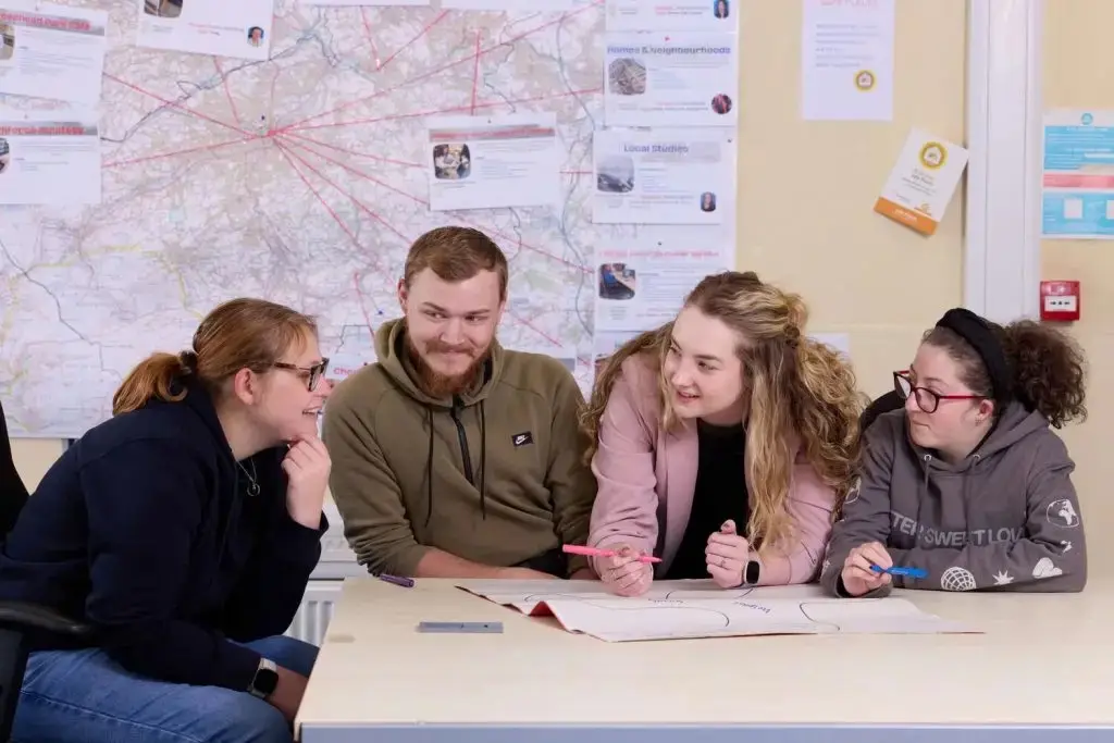 Four students around a table, smiling and discussing a map and notes in a college classroom setting.