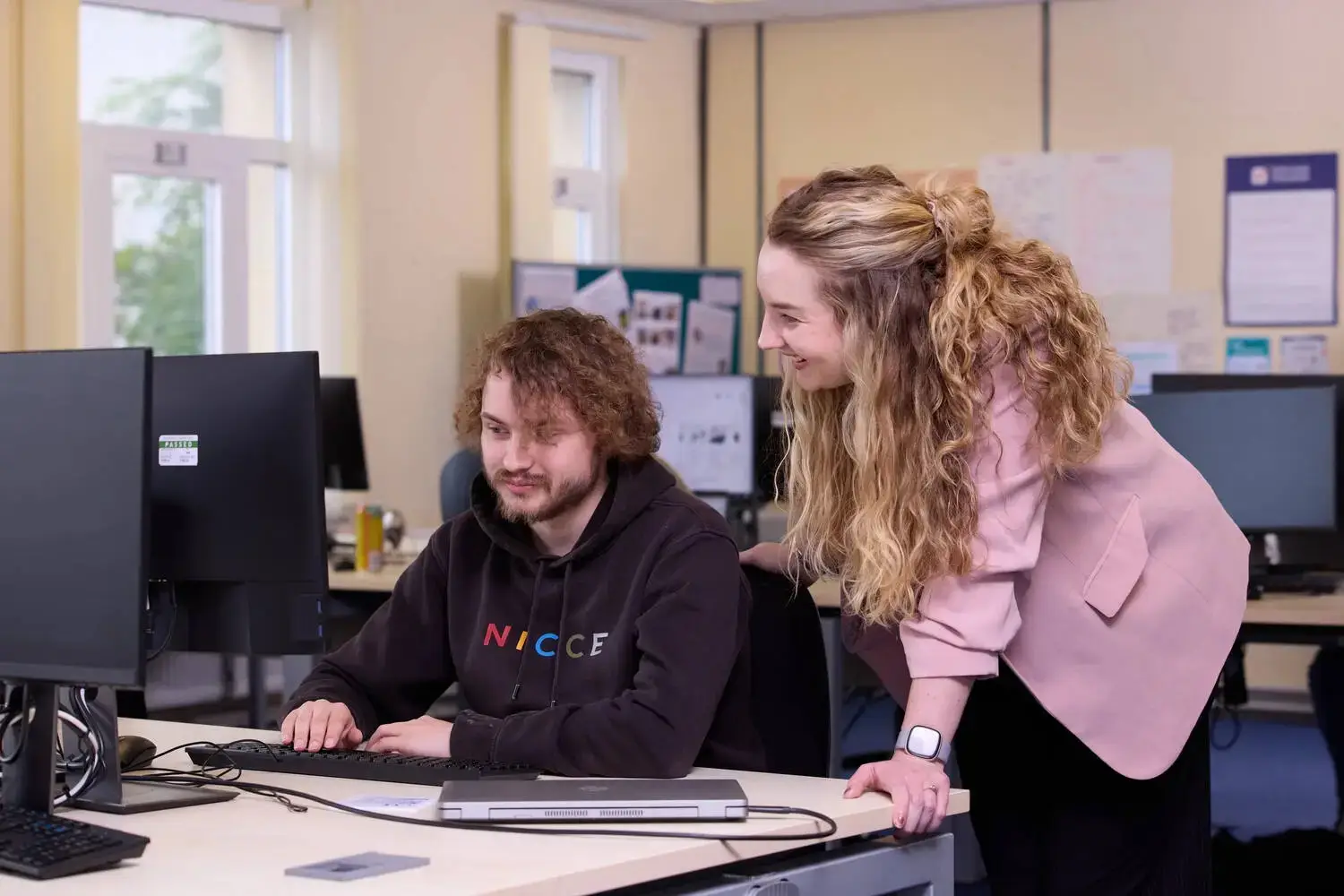 Two people in a college computer lab, one seated at a desk typing and the other standing, leaning in to observe the screen.