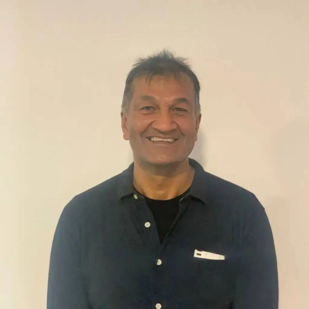 A man in a dark blue shirt smiling against a white background. He has short, dark hair and is wearing a black shirt underneath.