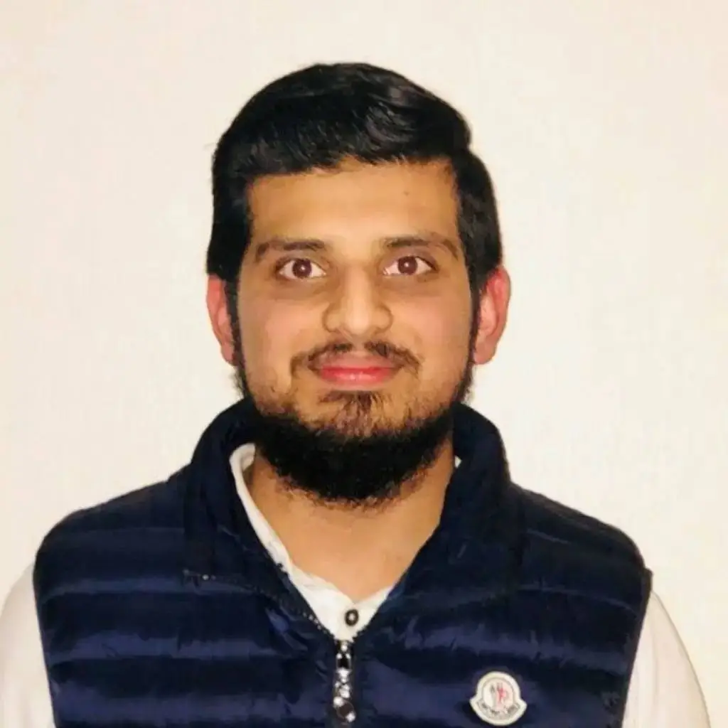A young man in a navy blue puffer vest and white shirt poses for a portrait against a white background.