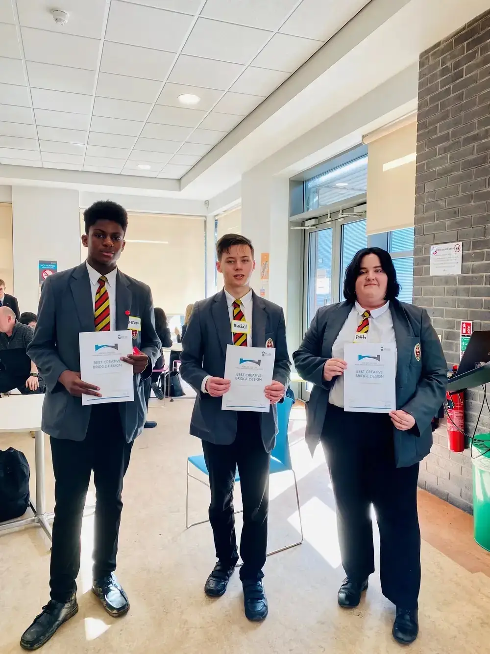 Three students in school uniforms holding certificates for 'Best Creative Bridge Design' in a college hallway.