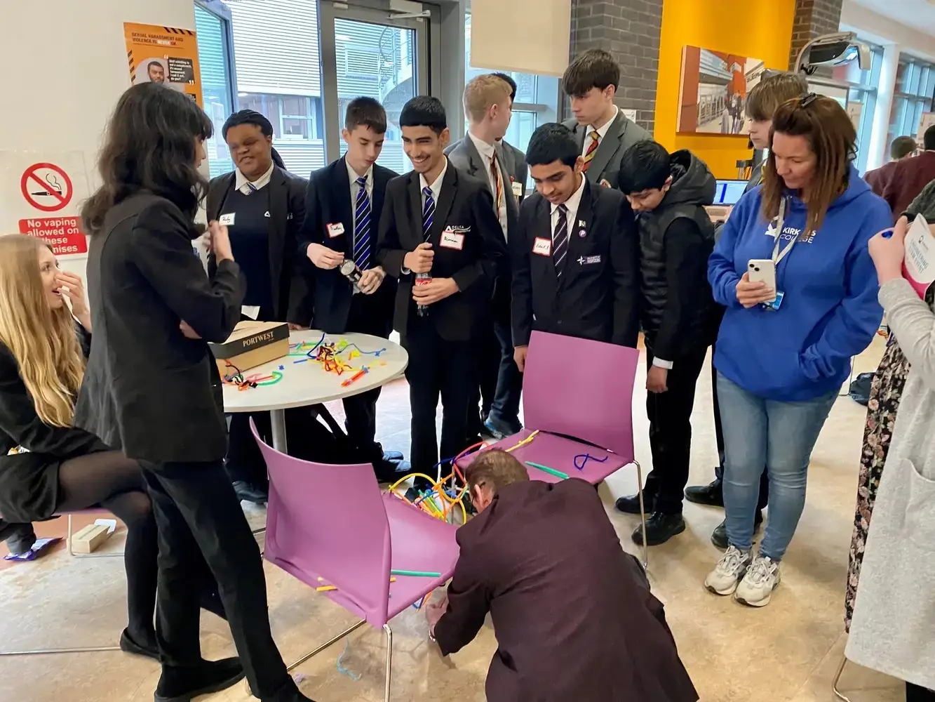 Students in school uniforms gather around a table with colourful engineering models, engaging in a hands-on learning activity with a presenter.