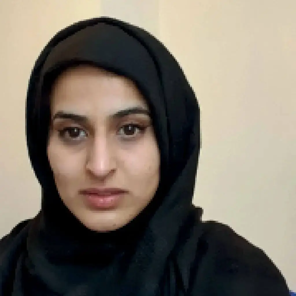 A woman in a black headscarf poses for a photo against a beige wall. She has brown eyes and is looking directly at the camera.