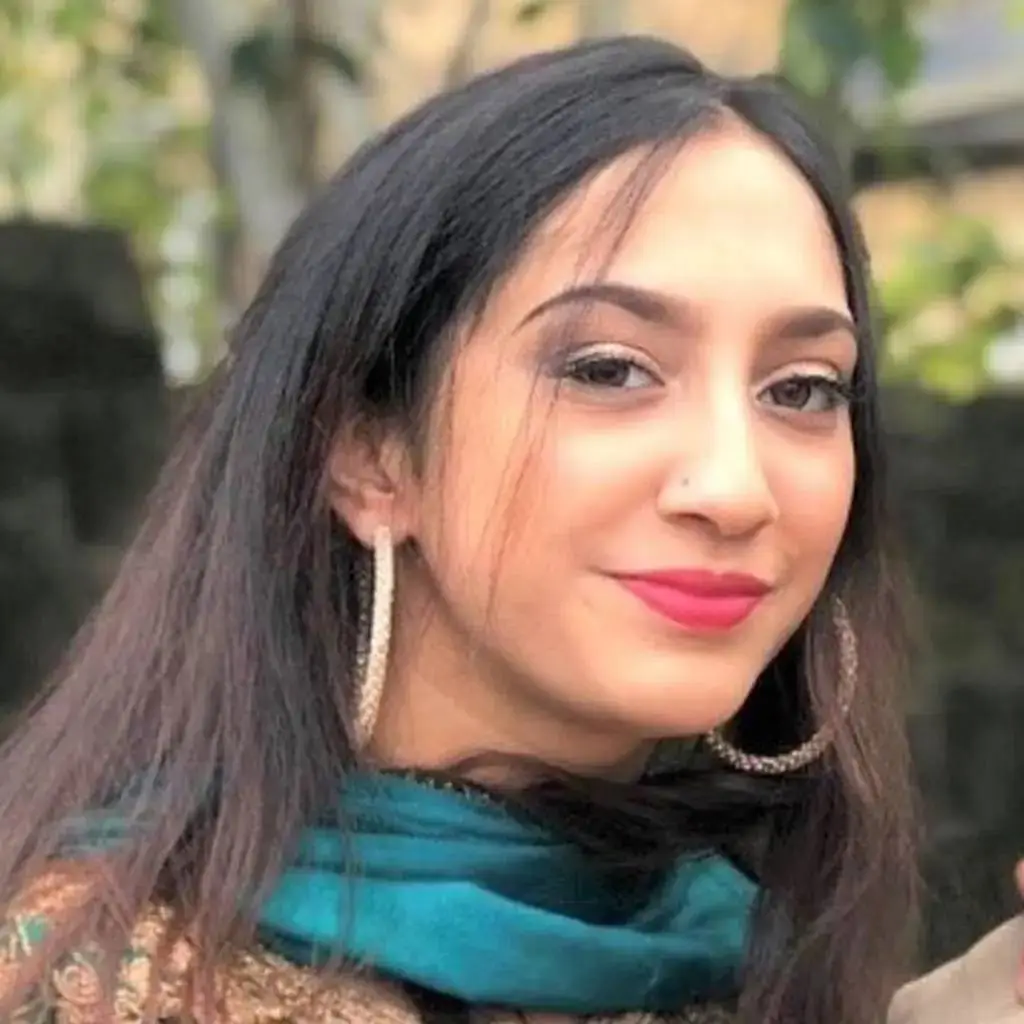 A woman with long dark hair and bright pink lipstick smiles, wearing a teal scarf and gold earrings. She stands outdoors with blurred greenery in the background.