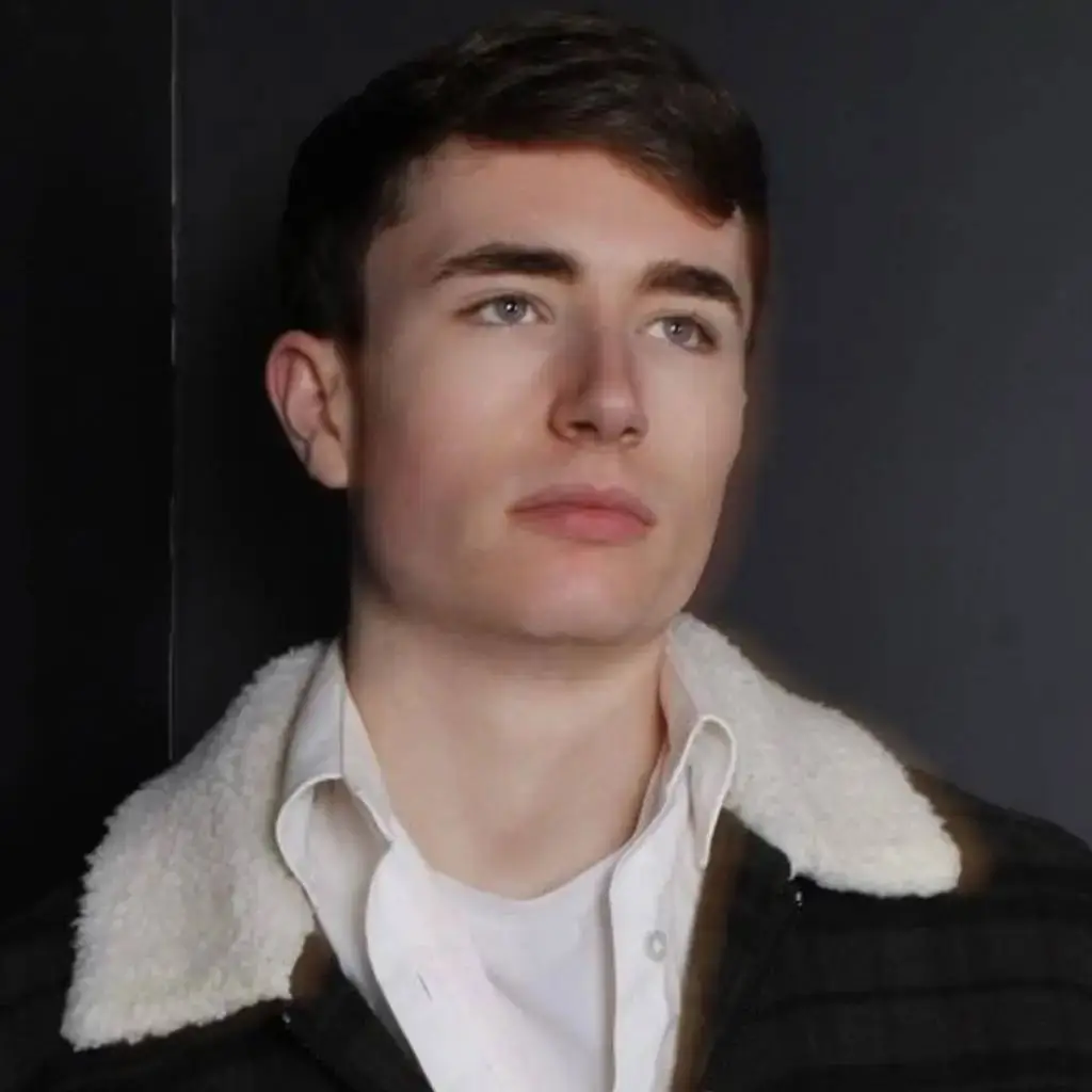 A young man in a white shirt and black jacket with a white collar poses for a portrait against a dark background.