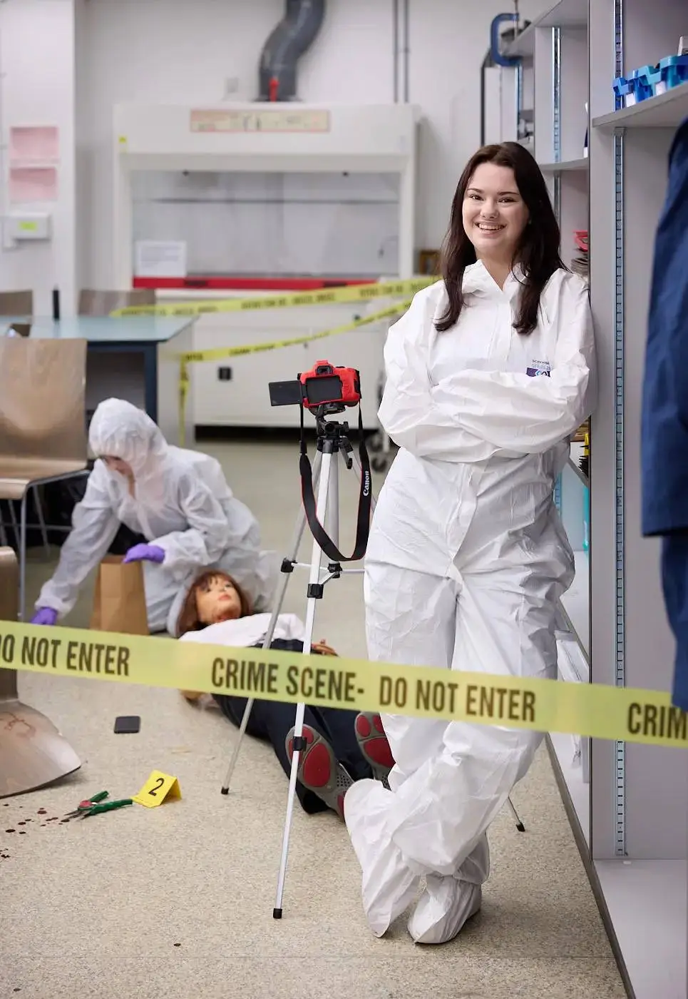 Two forensic science students in white protective suits at a crime scene mock-up, one posing with a camera on a tripod, the other examining evidence.