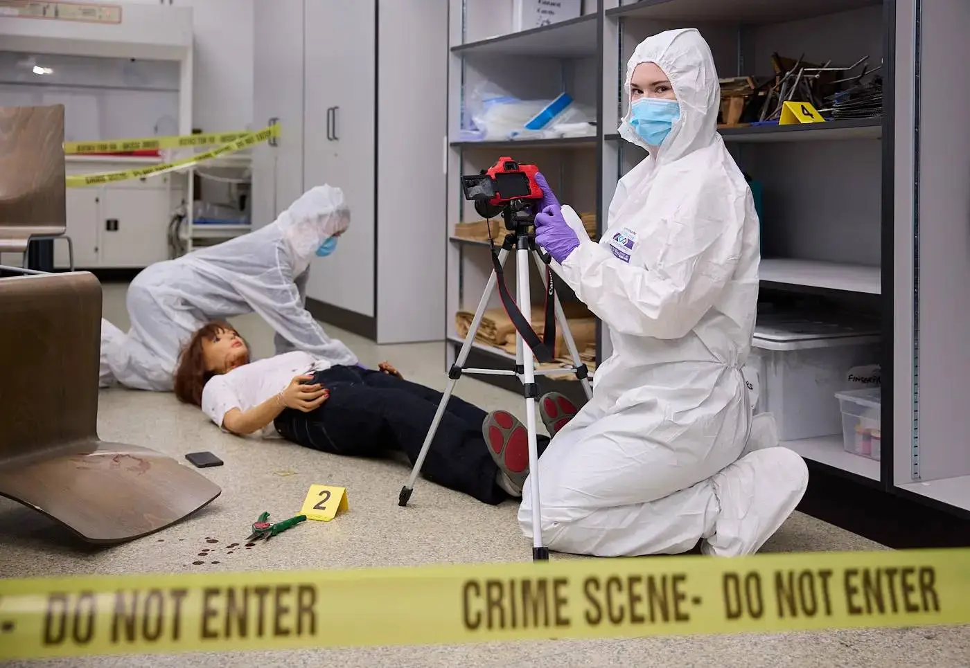 Two forensic scientists in white protective suits photograph a crime scene with a body and evidence markers in a laboratory setting.