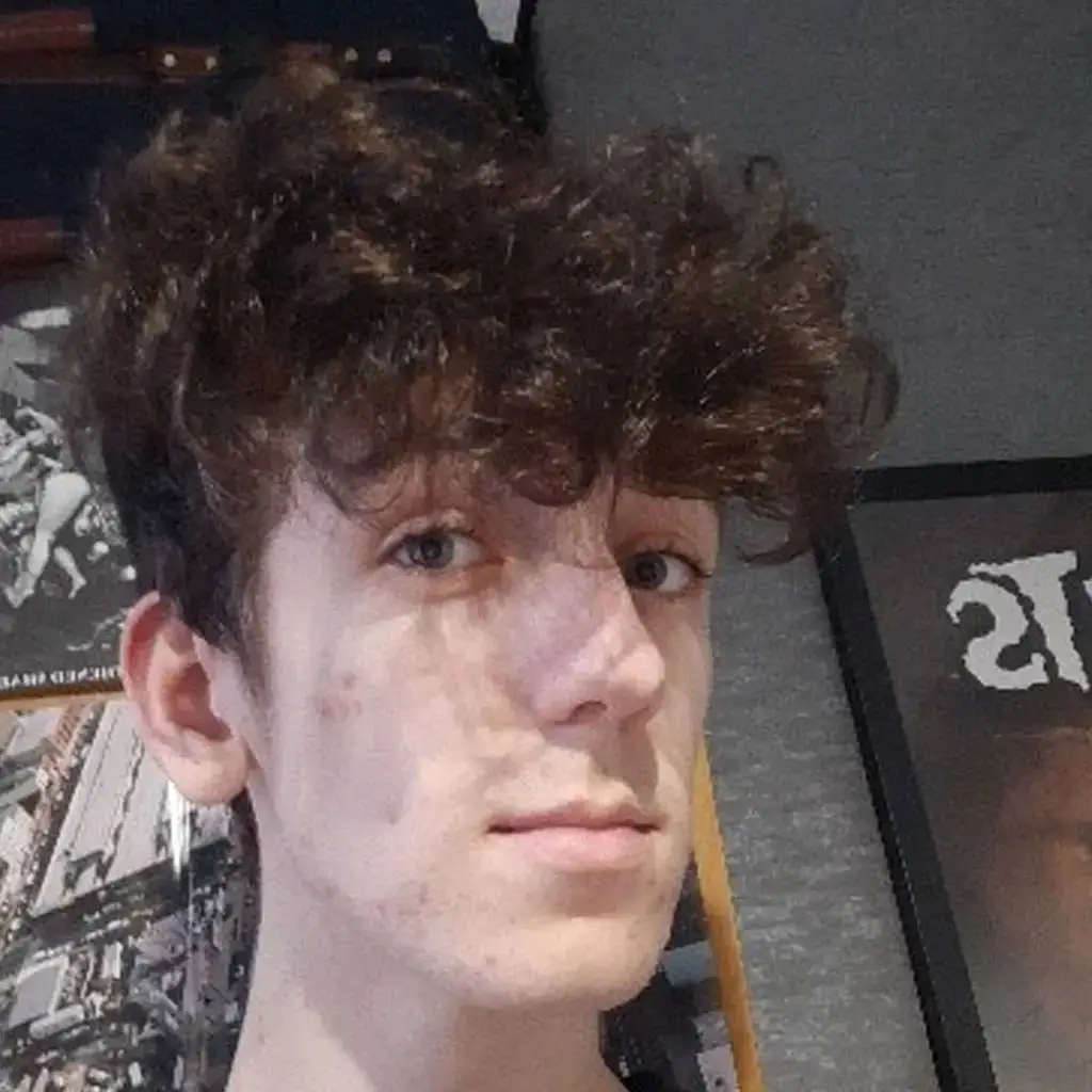 A young man with curly hair and a nose ring poses for a photo in front of a black and white poster.