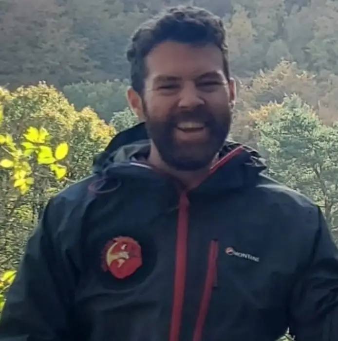 A man in a black jacket with a red zip and a red badge stands outdoors, smiling. Trees with yellow leaves are in the background.