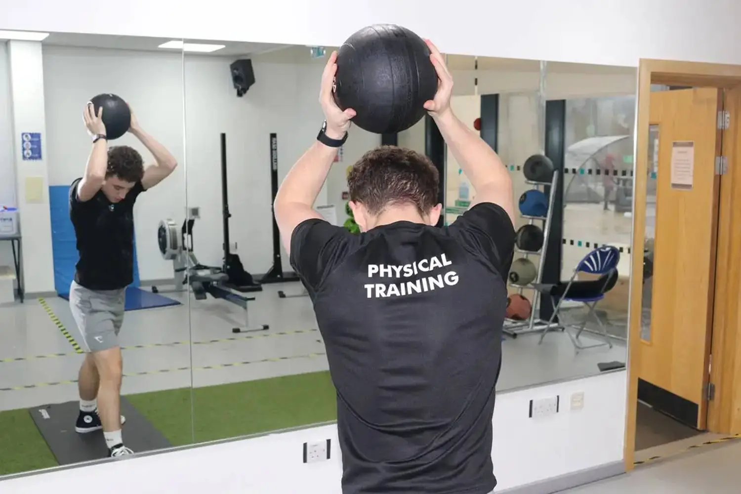 Two students in a gym perform overhead medicine ball exercises, reflecting in a mirror, wearing 'PHYSICAL TRAINING' shirts. Equipment and a door are visible in the background.