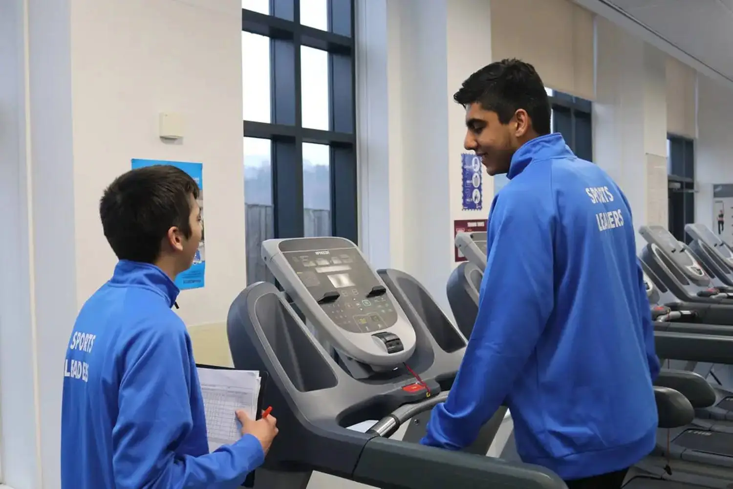 Two individuals in blue sports jackets, one on a treadmill and the other holding a clipboard, engaged in a discussion in a college gym.