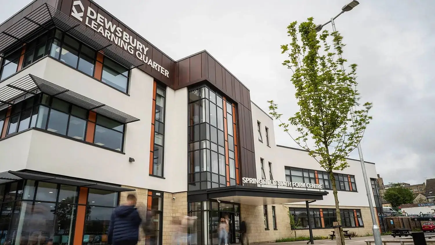 Modern college building with glass facade and signage. Students walking in front under a cloudy sky.