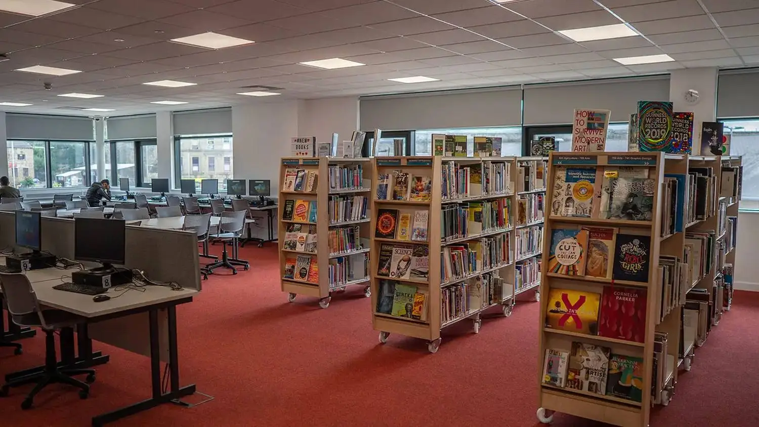A college library with bookshelves, computers, and red carpet. Large windows let in natural light.